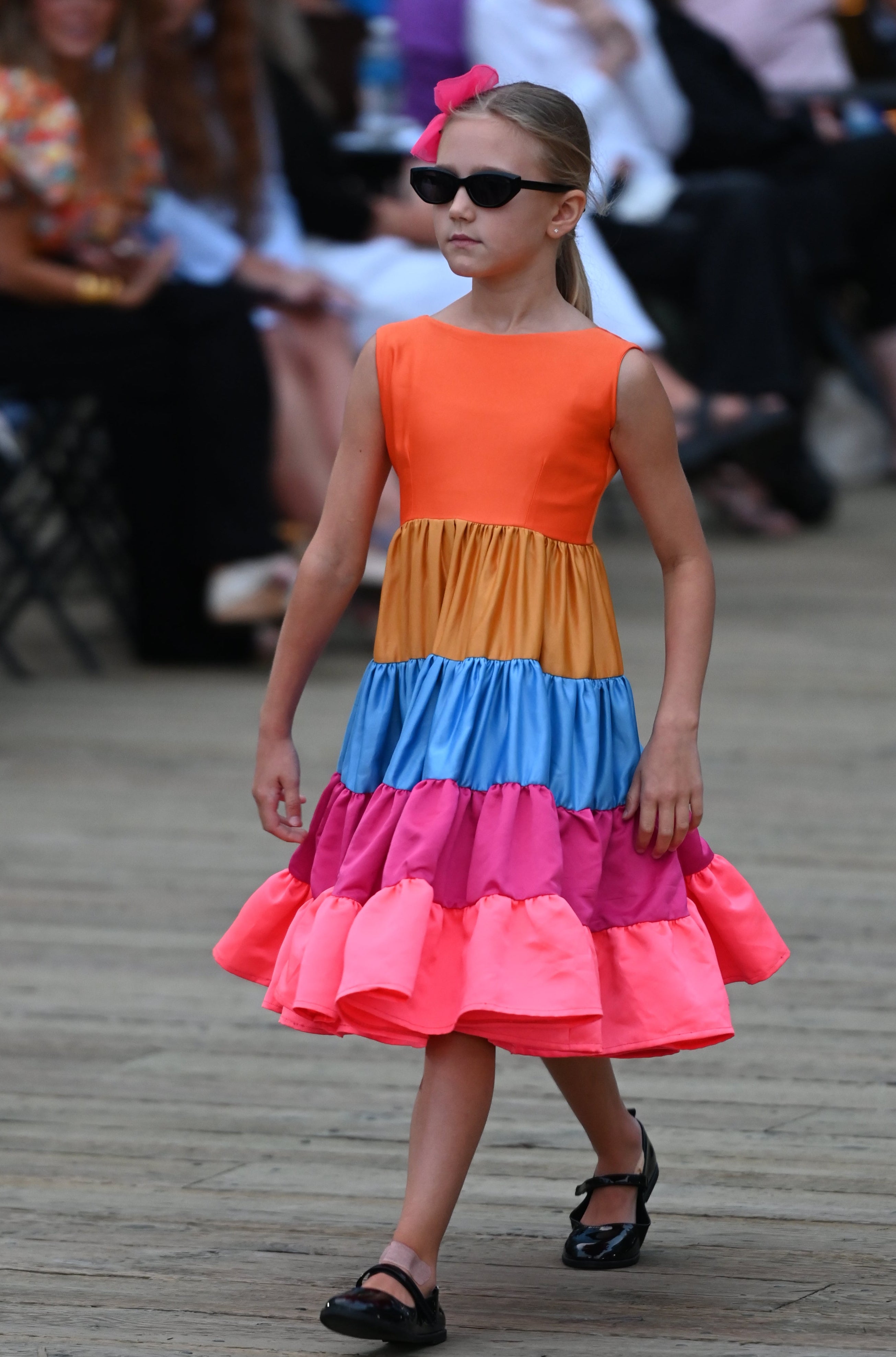 A young girl walking on a runway wearing a sleeveless, tiered, vibrant color-blocked dress with a round neckline and cinched waist.