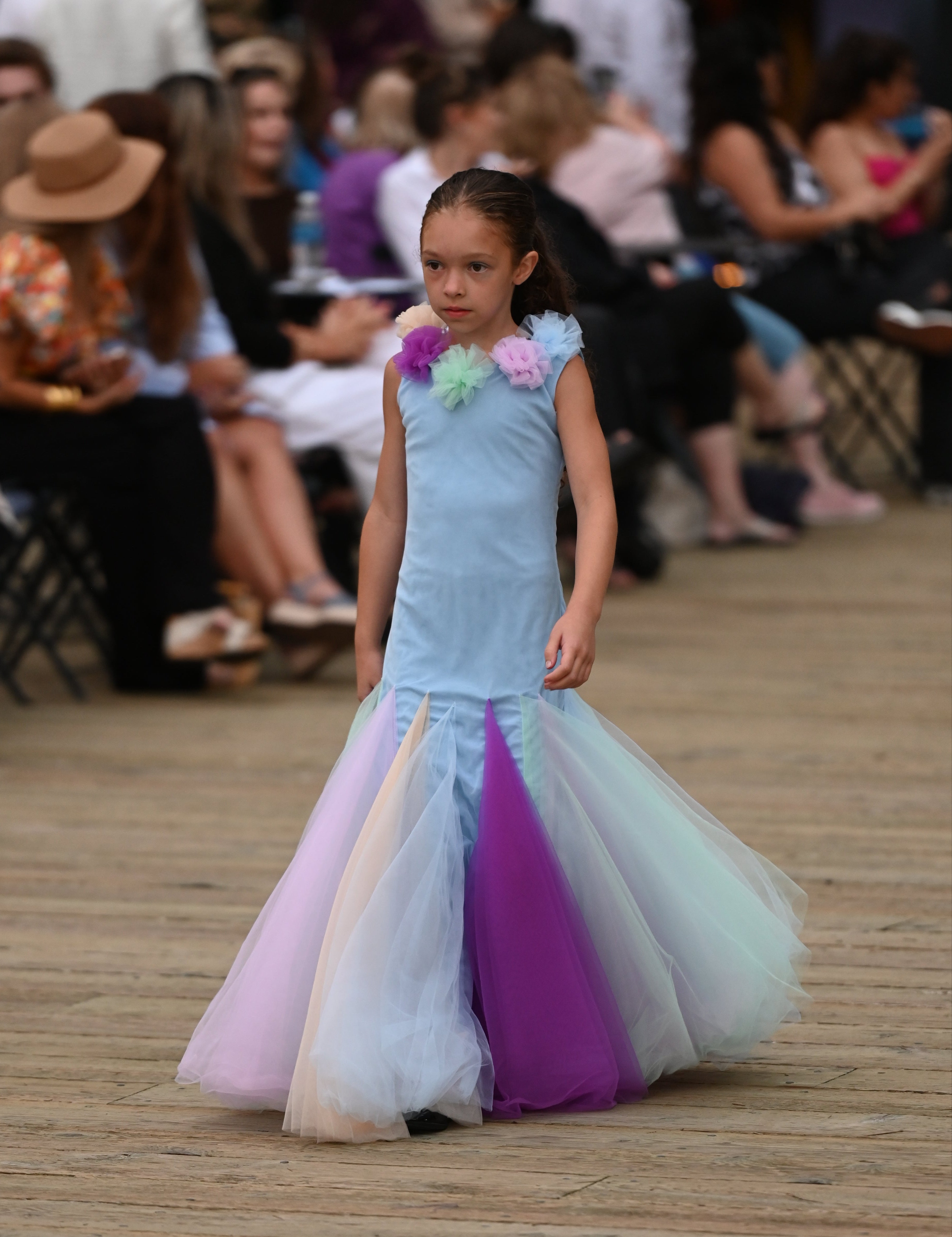 Young girl in a colorful dimazbrand dress standing on a wooden floor with a blurred crowd in the background